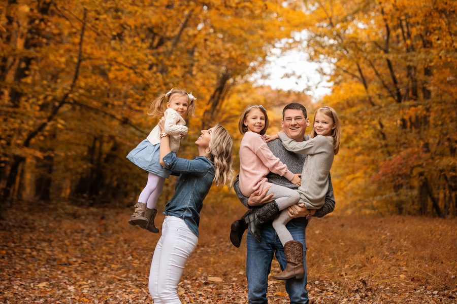 Parents lifting their two daughters, laughing, beneath golden autumn trees
