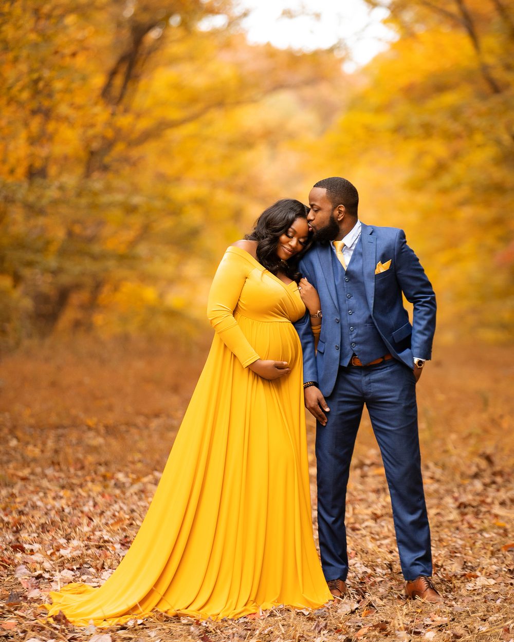 An expecting couple standing close together on a leaf-covered path